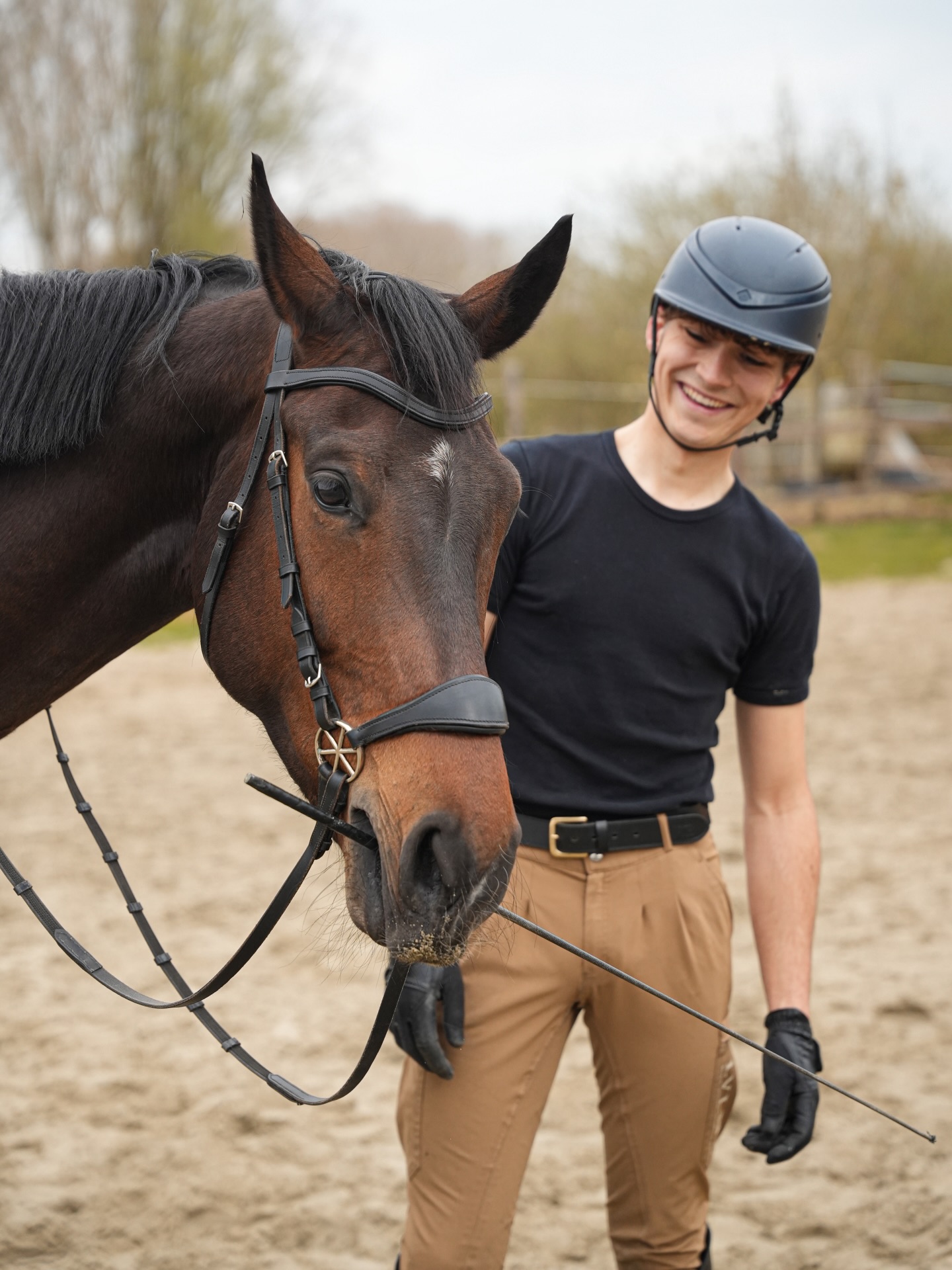 Robin Cooijmans lacht naar een jong bruin paard dat aan zijn zweep snuffelt in de buitenbak